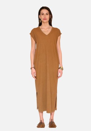 Femme debout face à nous, portant une robe midi marron sans manches avec des fentes latérales, des mocassins marron et des bijoux superposés.