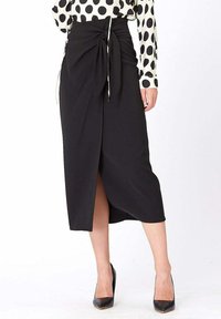 Jupe midi noire avec un design enroulé, présentant une fente avant et un système de fermeture par lien. Le tissu lisse et la taille ajustée accentuent la silhouette.