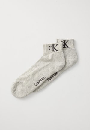 Chaussettes de cheville gris clair avec des bords côtelés, présentant un grand logo "cK" ; le texte "Calvin Klein" imprimé sur les côtés.