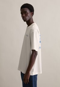 Relaxte pasvorm, off-white katoenen T-shirt met korte mouwen. Heeft blauwe grafische tekst op de achterkant, ronde halslijn en zijsplitten aan de zoom.