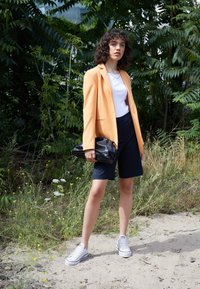 Mujer con cabello rizado que lleva un blazer amarillo, una blusa blanca, pantalones cortos azul marino, deportivas blancas y sostiene un clutch negro texturizado al aire libre, junto a la vegetación.