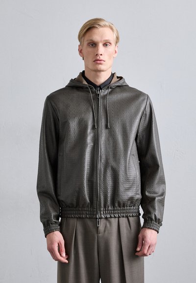 Hooded bunda vyrobená z texturovaného tmavě šedého materiálu s celoplošným vzorem. Má přední zip, elastické manžety a lem. V kombinaci s kalhotami na míru.
