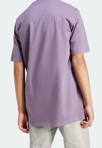 Person som bär en löst sittande, kortärmad lavendelfärgad t-shirt och ljusbeiga byxor, sedd bakifrån mot en enkel bakgrund.