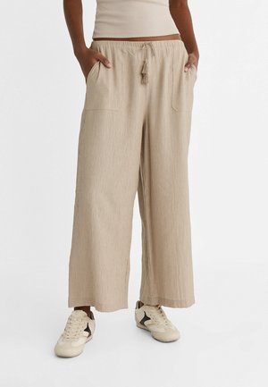 Vrouw draagt beige wijde broek met klepzakken en een trekkoord met kwastjes, gecombineerd met beige en zwarte sneakers.