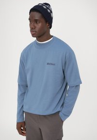 Svijetloplava dukserica kratkih rukava i opuštenog kroja, preko bijele košulje dugih rukava. Ima mali logotip na prsima.