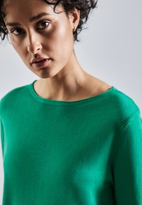 Pull vert avec un col large, texture douce et coupe ajustée. La vue en gros plan met en valeur la surface lisse du tissu et la profondeur de la couleur.