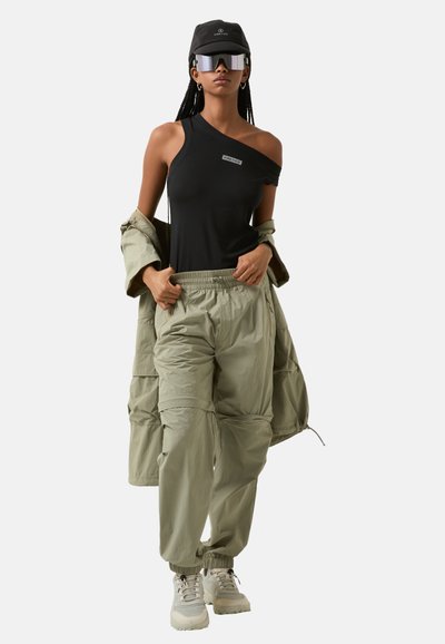 Černý top na jedno rameno, olivové cargo kalhoty s elastickými manžetami, oversized béžová bunda. Doplňky zahrnují černou kšiltovku a zrcadlové sluneční brýle.