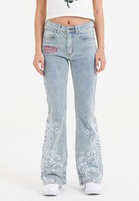 Kvinde i en hvid crop top og flagrende lyseblå jeans med intrikate hvide broderier på benene, iført hvide sneakers, stående foran en hvid baggrund.