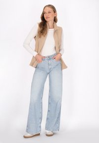 Beige ärmellose Weste über einem weißen Langarmshirt, kombiniert mit hellblauen, hoch taillierten Weitbeinjeans und weißen Sneakern.