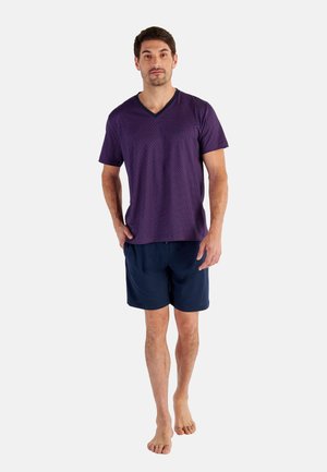 Mann steht barfuß, trägt ein violettes kurzärmeliges V-Ausschnitt-Shirt und marineblaue Shorts vor einem einfarbigen Hintergrund.