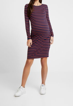 Pregnant woman wearing a navy blue and red striped knee-length dress with long sleeves and white sneakers, standing against a plain background.