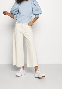 Top de manga abullonada de color azul claro con cuello redondo, combinado con pantalones de pierna ancha de talle alto en color crema y zapatillas blancas.