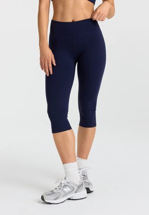 Kvinde iført marineblå korte leggings, hvide sokker og grå sports sko, stående mod en lys baggrund.
