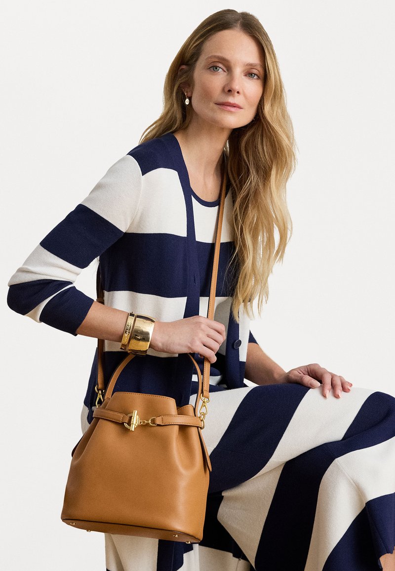 Brązowa skórzana torba typu bucket z ściąganym zamkiem, złotymi akcentami i odpinanym paskiem na ramię. Charakteryzuje się gładką fakturą i minimalistycznym designem.