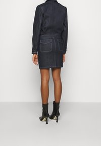 Mörk denimjacka klänning med en åtsittande silhuett, kontrasterande blå sömmar, två bakfickor och en bälte vid midjan, i kombination med svarta ankelstövlar.