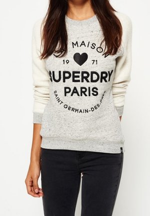 Femme portant un pull gris clair avec des manches crème et un texte noir indiquant "Maison Superdry Paris 1971 Saint Germain-Des-Prés."
