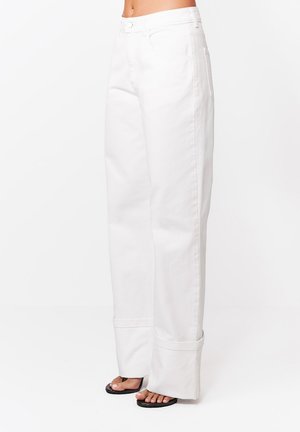 Białe spodnie z wysokim stanem i szerokimi nogawkami z podwiniętymi mankietami, noszone z czarnymi sandałami na obcasie z odkrytymi palcami, widoczne od talii do stóp.