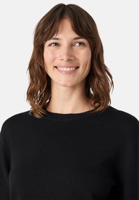 Černý, strukturovaný pletený svetr s kulatým výstřihem, který má volný střih a jemné detaily prošití. Modelka má vlasy po ramena.