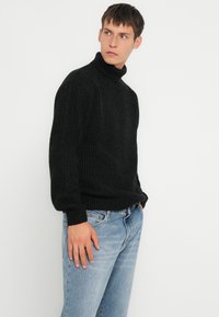 Svart ribbestrikket topp med høy hals, avslappet passform og lange ermer, kombinert med lyseblå jeans. Myk tekstur og sømløst design.