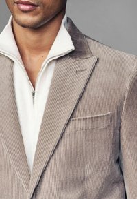 Beige Cordblazer mit einem Reverskragen und einer Brusttasche, kombiniert mit einem weißen Zip-Hoodie aus sanftem Material.