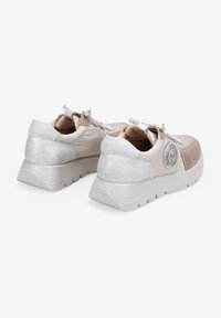 Zapatillas beige con parte superior texturizada, acentos plateados en el talón y suela blanca gruesa. Incluye un detalle de logo circular en el lado.