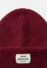 Bordo pletena kapa s rebrastom teksturom, s bijelom etiketom na kojoj piše "MADS NØRGAARD COPENHAGEN" na prednjoj strani. Mekani akrilni materijal.