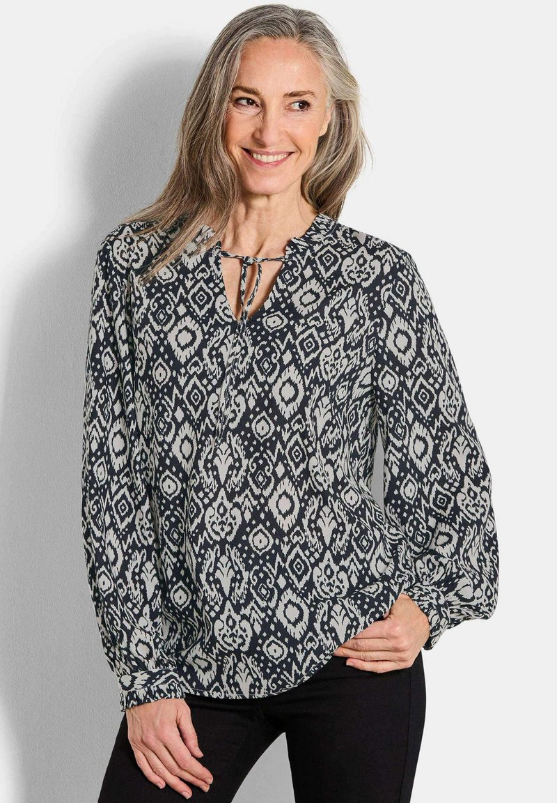 Fließende, gemusterte Bluse in Schwarz und Creme mit einer Schlaufe am Halsausschnitt. Merkmale sind lange Ärmel und eine entspannte Passform mit elastischen Bündchen.