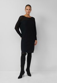 Femme portant une robe en tricot côtelé noire avec des manches longues transparentes et des bottes noires à talons hauts au-dessus du genou, se tenant contre un mur clair et uni.