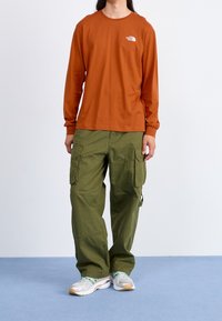 Pomarańczowa bawełniana koszula z długim rękawem z białym logo; zielone spodnie cargo z dużymi kieszeniami; szare trampki z zielonymi akcentami i teksturowaną podeszwą.