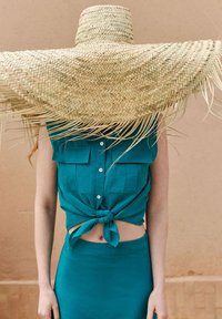 Top corto in lino color teal con dettagli di bottoni e tasche, abbinato a una gonna fitted coordinata. Un grande cappello di paglia oscura il volto.