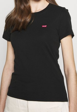 Femme portant un t-shirt noir ajusté Levi’s avec un patch logo rouge sur la poitrine et un pantalon clair sur un fond clair.
