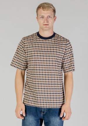 Blonder Mann mit kurzärmligem beige-braun-navy kariertem Hemd mit dunklem Kragen und blauer Jeans, vor einfarbigem Hintergrund stehend.