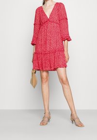 Una donna sta in piedi indossando un vestito rosa con stampa leopardo e volants, tiene in mano una piccola borsa beige e indossa sandali con tacco beige e cinturini.