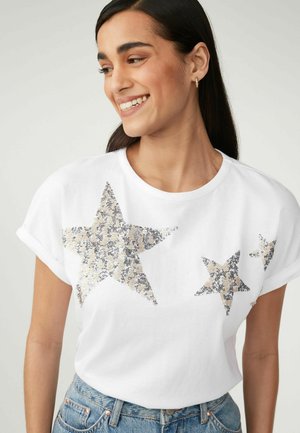 T-shirt blanc à manches courtes retroussées, arborant trois grands motifs d'étoiles en sequins métalliques dans des tons argentés et beiges.