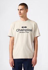 Hombre con el cabello corto que lleva una camiseta beige de Champion con el texto "Only for Members", mirando hacia la izquierda contra un fondo blanco liso.