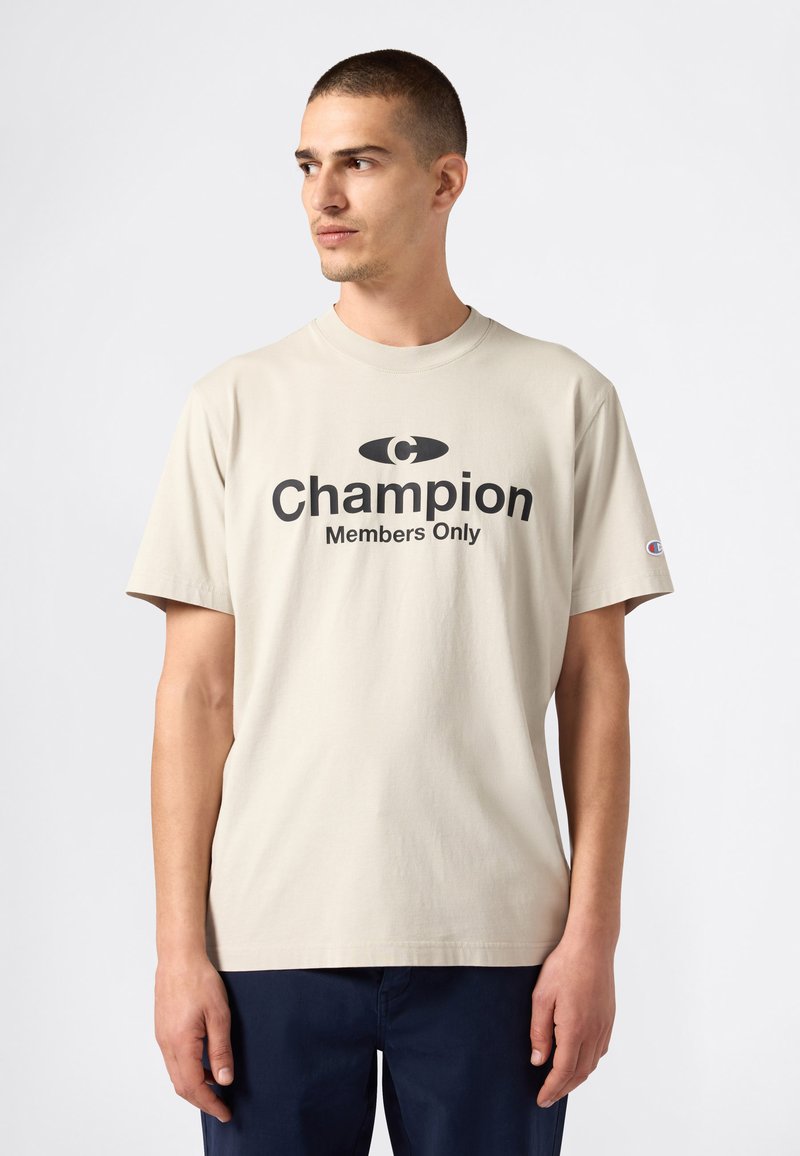 Hombre con el cabello corto que lleva una camiseta beige de Champion con el texto "Only for Members", mirando hacia la izquierda contra un fondo blanco liso.