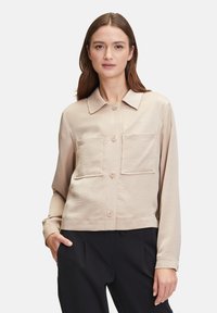 Beige cropped shirt met een knoopsluiting aan de voorkant, twee borstzakken en lange mouwen. Soepele textuur en een klassiek kraagontwerp.