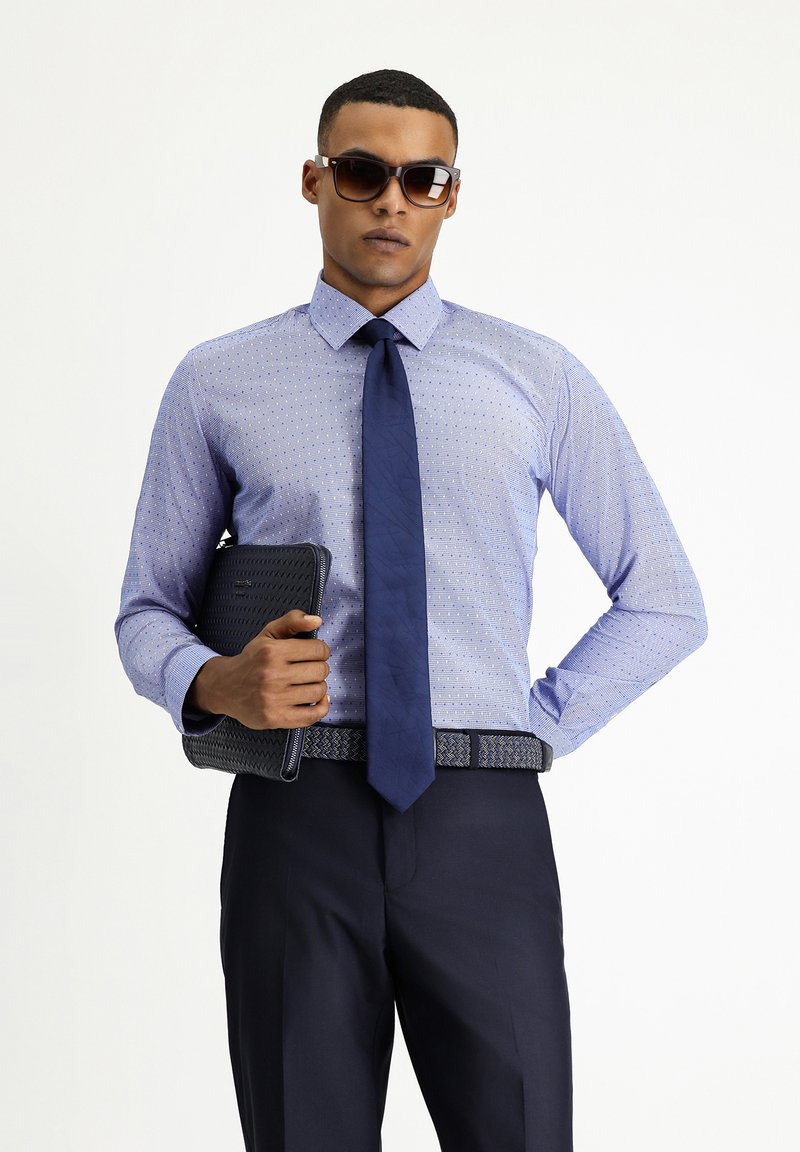 Lichtblauw gedrukt shirt met een gestructureerde kraag, gecombineerd met een marineblauwe stropdas, marineblauwe broek en een gestructureerde zwarte clutch.