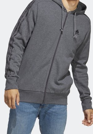 Hombre con sudadera con cremallera gris de Adidas con rayas negras en las mangas y vaqueros azul claro, mostrado desde el pecho hasta la cadera.