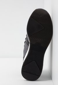Semelle d'une basket noire avec un motif texturé et le logo "Michael Kors" sur le talon, positionnée verticalement sur fond blanc.