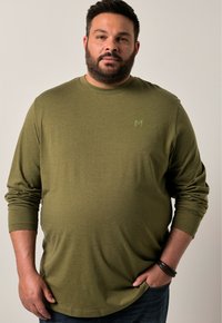 Langärmliges olivgrünes Shirt aus weichem Material mit rundem Halsausschnitt und kleinem Logo auf der Brust, kombiniert mit blauen Jeans.