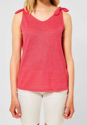 Kobieta w czerwonym bezrękawniku dzianym z wiązaniem na ramieniu i białych krótkich spodenkach do kolan, stojąca na jednolitym tle.