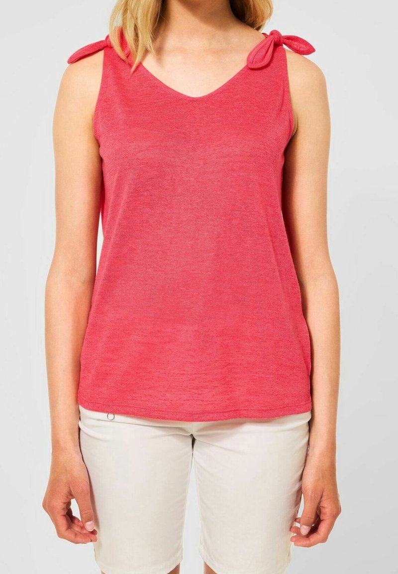 Femme portant un top en maille rouge sans manches avec un détail de nœud à l'épaule et un short blanc mi-genoux, debout devant un fond uni.