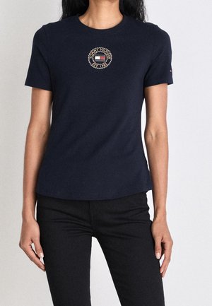 Kobieta ubrana w granatową koszulkę z krótkim rękawem z okrągłą naszywką z logo z napisem "Tommy Hilfiger Est. 1985" na piersi.