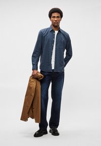 Μπλε καρό πουκάμισο με κουμπιά, λευκή μπλούζα από κάτω, σκούρα τζιν, κρατώντας ένα καφέ μπουφάν, μαύρα παπούτσια, στέκεται μπροστά σε ανοιχτό φόντο.