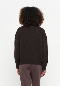Brązowy, oversize sweter z miękkiego materiału dzianinowego, z wysokim kołnierzem i prążkowanymi mankietami. Noszony z luźnymi, jasnobrązowymi spodniami.