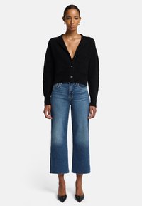 Cardigan nero corto con scollo a V e due tasche frontali, abbinato a jeans blu a vita alta e tacchi neri a punta.