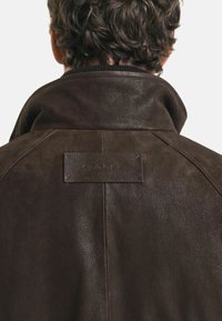 Bruine leren jas met een hoge kraag, textuur en een GANT logo patch op de achterkant. Eenvoudig en functioneel ontwerp.