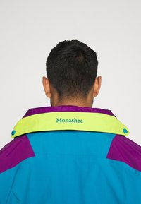 Homme portant une veste bleue, violette et jaune avec le texte «Monashee» sur le col jaune, vu de dos sur un fond uni.