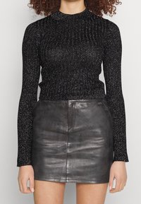 Kvinna som bär en svart glittrig långärmad topp instoppad i en svart läder minikjol, står mot en enkel bakgrund.